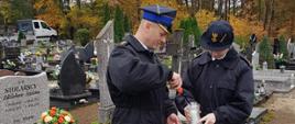 Zdjęcie przedstawia mężczyznę i kobietę w mundurach strażackich zapalających znicz. Stója przed grobem, oddając hołd zmarłym.
