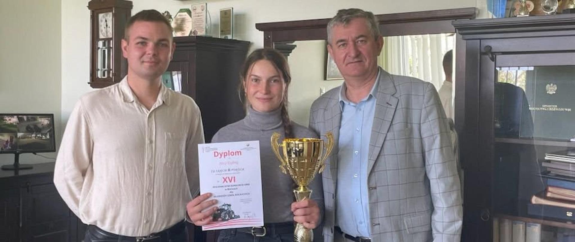 Pamiątkowe zdjęcie z uroczystego wręczenia nagród: w środku uczennica Alicja Szyling, laureatka II miejsca w konkursie sprawnościowym, dumnie prezentuje zdobyty dyplom oraz okazały złoty puchar. Obok niej stoją opiekun Bartosz Maćkowski (po lewej) oraz przedstawiciel dyrekcji szkoły (po prawej). W tle widoczne są liczne trofea i puchary zgromadzone na półkach w szkolnym gabinecie.