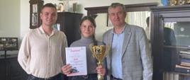 Pamiątkowe zdjęcie z uroczystego wręczenia nagród: w środku uczennica Alicja Szyling, laureatka II miejsca w konkursie sprawnościowym, dumnie prezentuje zdobyty dyplom oraz okazały złoty puchar. Obok niej stoją opiekun Bartosz Maćkowski (po lewej) oraz przedstawiciel dyrekcji szkoły (po prawej). W tle widoczne są liczne trofea i puchary zgromadzone na półkach w szkolnym gabinecie.