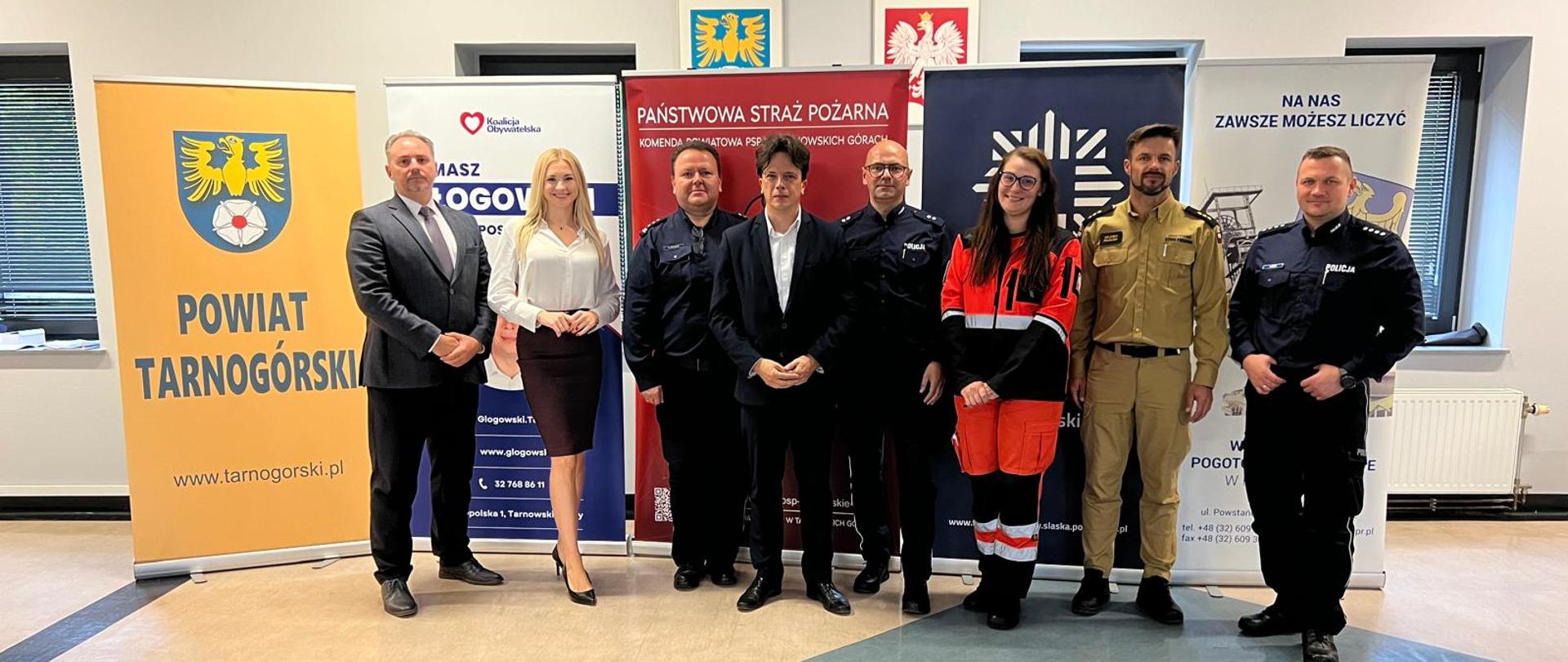 W jednym rzędzie soją od lewej: mężczyzna w garniturze, kobieta, policjant, mężczyzna w garniturze, policjant, pani ratownik medyczny, strażak i policjant
