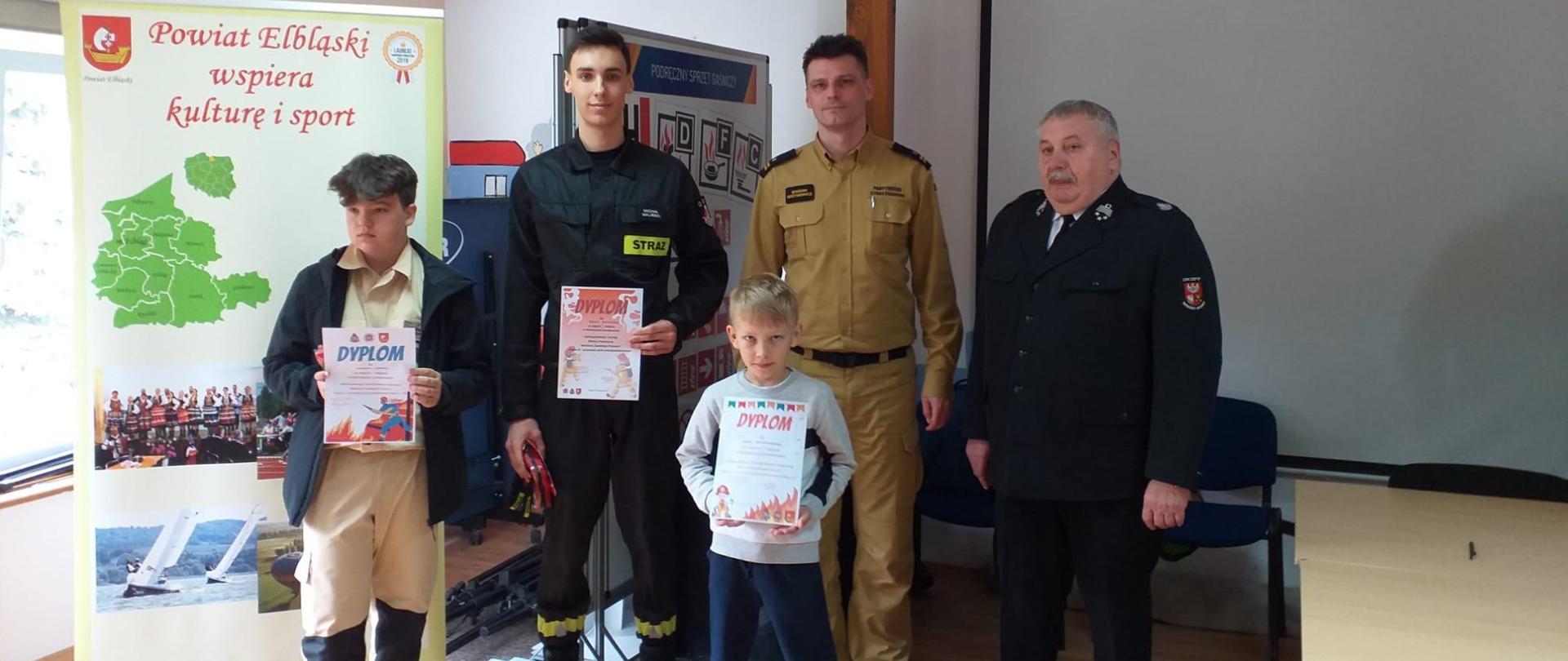 Na zdjęciu troje uczestników dumnie pokazuje swoje dyplomy, obok nich znajduje się Komendant Miejski i Druh Prezes Związku Ochotniczej Straży Pożarnej. 