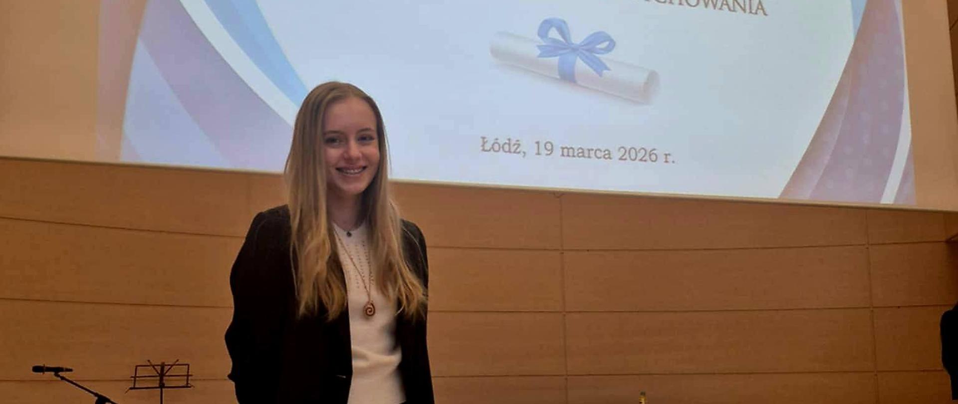 Zdjęcie: dziewczyna w galowym stroju stoi na tle banera Stypendium Prezesa Rady Ministrów 