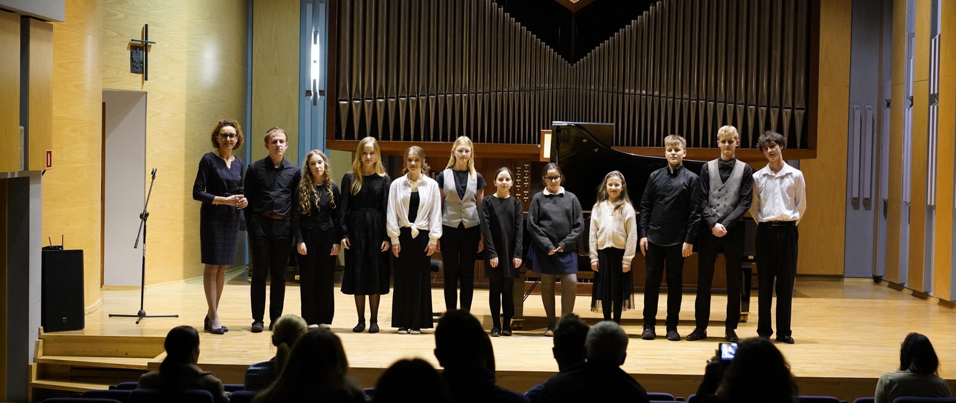 Zdjęcie przedstawia grupę jedenastu młodych artystów oraz ich opiekunkę stojących w rzędzie na scenie sali koncertowej, kłaniając się lub prezentując po występie. Po lewej stronie stoi dorosła kobieta w ciemnej, eleganckiej sukni. Obok niej znajduje się grupa dzieci i młodzieży w różnym wieku, ubranych w stroje galowe (dominują kolory czarny i biały, kamizelki oraz koszule). Centralnym punktem tła są monumentalne organy piszczałkowe o charakterystycznym, symetrycznym układzie rur przypominającym literę „V”. Po prawej stronie, za plecami artystów, widoczny jest fragment czarnego, lśniącego fortepianu skrzydłowego. Perspektywa: Zdjęcie zostało wykonane z poziomu widowni; w dolnej części kadru widoczne są ciemne sylwetki widzów siedzących w fotelach, co podkreśla relację między wykonawcami a publicznością. Otoczenie: Sala koncertowa ma nowoczesny charakter z jasnymi, drewnianymi ścianami bocznymi i profesjonalnym oświetleniem punktowym skierowanym na artystów.