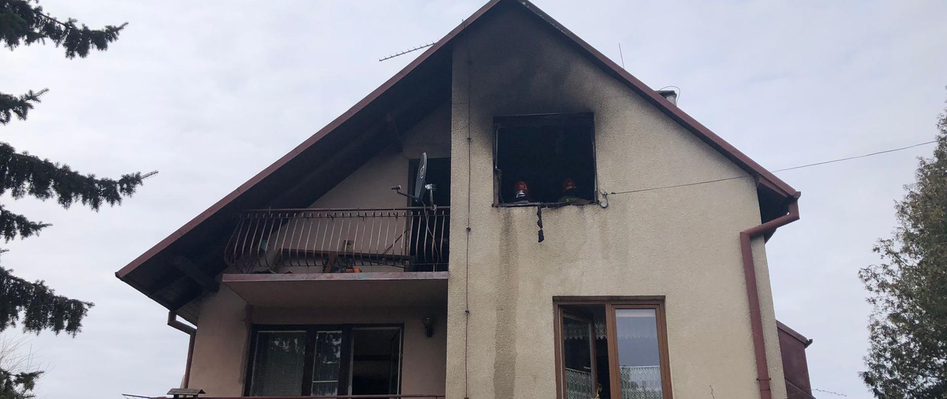 Zdjęcie przedstawia cały budynek od frontu. Budynek murowany, pokryty czerwoną blachą. Lewa strona budynku koloru pomarańczowego, natomiast prawa część szaro – żółtego. Na najwyższej kondygnacji widać otwór okienny, a nad nim okopconą na czarno ścianę budynku. W pomieszczeniu, w którym występował pożar, widać dwóch strażaków ubranych w ubranie specjalne typu Nomex wraz z czerwonymi hełmami na głowach. Na zdjęciu widać okna z brązowymi futrynami, które są otwarte, w celu oddymienia budynku.