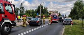 na drodze po lewej stronie stoi czarny samochód na środku stoi karetka a po prawej stronie drugi samochód z wgniecionym tyłem przy samochodzie stoją strażacy ratownik medyczny a na chodniku stoi policjant i rozmawia z mężczyzną i kobietą