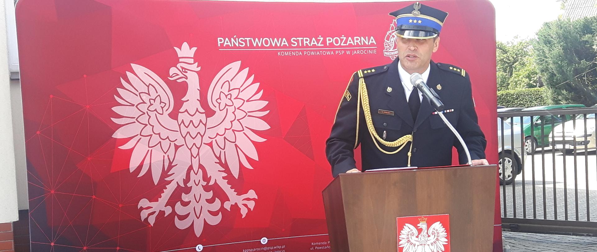 strażak w rogatywce i mundurze ze sznurem sto przed mównicą, przemawia, w tle czerwona ścianka tekstylna z orlem