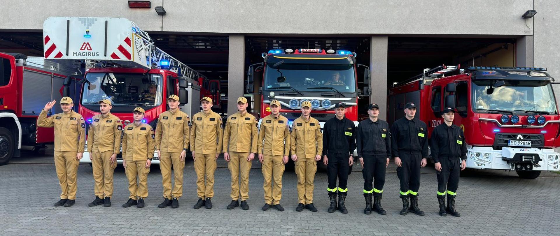 Strażacy stoją przed jednostką, oddając hołd, za nimi samochody ratowniczo-gaśnicze z włączoną sygnalizacją świetlną