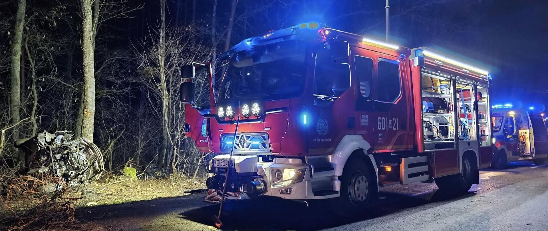 Pionowe, nocne zdjęcie przedstawia czerwony wóz strażacki z włączonymi światłami awaryjnymi, stojący na poboczu drogi, prawdopodobnie na miejscu zdarzenia.
Wóz strażacki, znajdujący się w centrum kadru, jest czerwony z białymi elementami. Na jego dachu i z przodu kabiny świecą niebieskie światła sygnalizacyjne. Na boku pojazdu widoczny jest numer "601-21". Przedni zderzak jest wyposażony w dodatkowe oświetlenie. Z tyłu wozu, częściowo widoczny, znajduje się kolejny pojazd ratunkowy, karetka pogotowia, również z włączonymi niebieskimi światłami.
Po lewej stronie zdjęcia, na tle ciemnych drzew pozbawionych liści, widoczne są fragmenty rozbitego pojazdu, który brał udział w wypadku drogowym. Ciemność nocy jest rozświetlona przez światła pojazdów ratunkowych oraz maszt oświetleniowy umieszczony na dachu samochodu ratowniczo - gaśniczego. Droga, na której stoją pojazdy, jest asfaltowa i mokra, co może wskazywać na niedawne opady deszczu. Ogólna atmosfera zdjęcia jest poważna i przedstawia interwencję służb ratunkowych.
