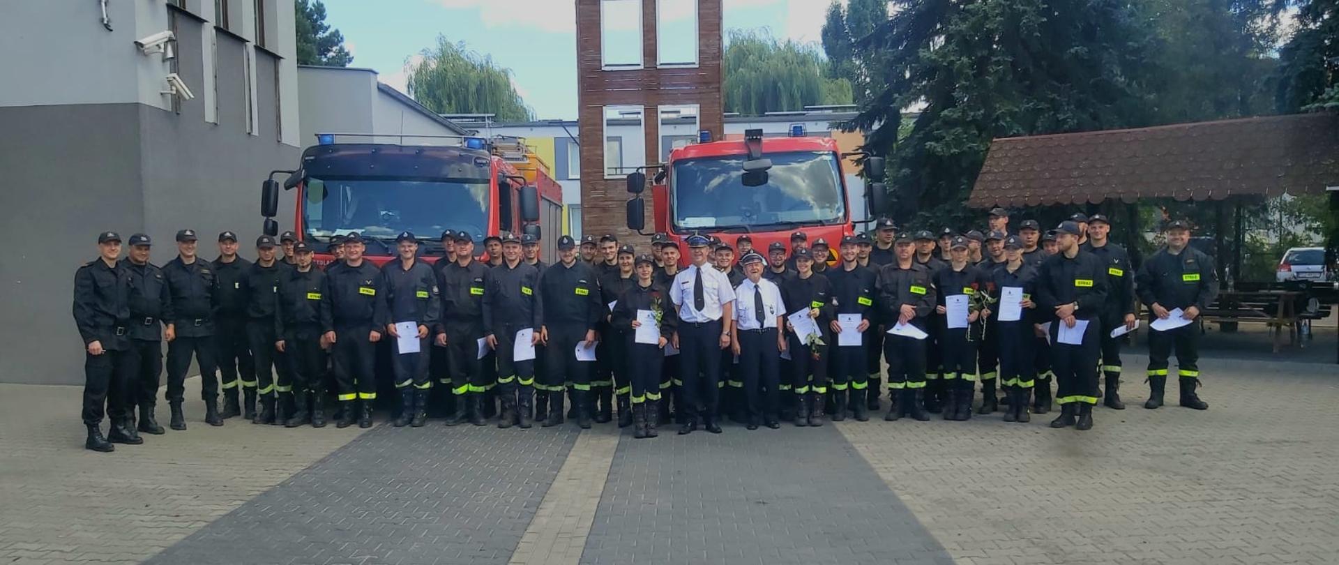 Strażacy uczestnicy kursu, wraz Komendantem Powiatowym i kadrą dydaktyczną po wręczeniu dyplomów ukończenia kursu, na tle czerwonych samochodów bojowych i budynki Komendy w Szamotułach
