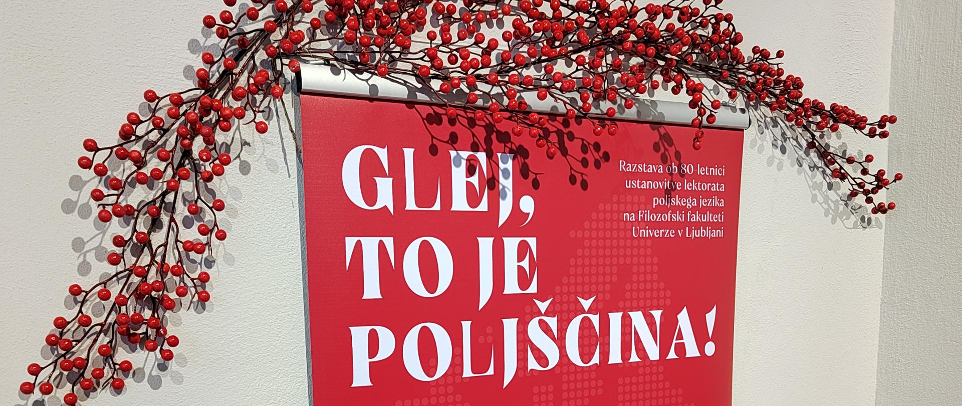 Czerwona plansza tytułowa wystawy „Glej, to je poljščina!” ustawiona w sali wystawowej. Planszę zdobi biały motyw kwiatowego serca, a nad nią znajduje się dekoracja z gałązek z czerwonymi owocami. Na stole poniżej leżą materiały informacyjne i edukacyjne o Polsce.
