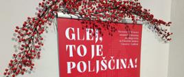 Czerwona plansza tytułowa wystawy „Glej, to je poljščina!” ustawiona w sali wystawowej. Planszę zdobi biały motyw kwiatowego serca, a nad nią znajduje się dekoracja z gałązek z czerwonymi owocami. Na stole poniżej leżą materiały informacyjne i edukacyjne o Polsce.