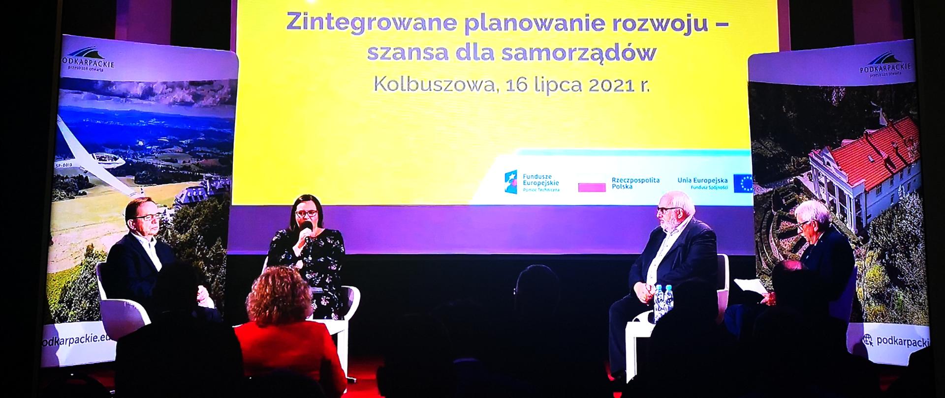 Cztery osoby (trzech mężczyzn i kobieta - minister Małgorzata Jarosińska-Jedynak) siedzą na scenie. Przed nimi zarys ludzi na widowni. Za nimi ekran z napisem Zintegrowane planowanie rozwoju - szansa dla samorządów. Kolbuszowa, 16 lipca 2021 r.