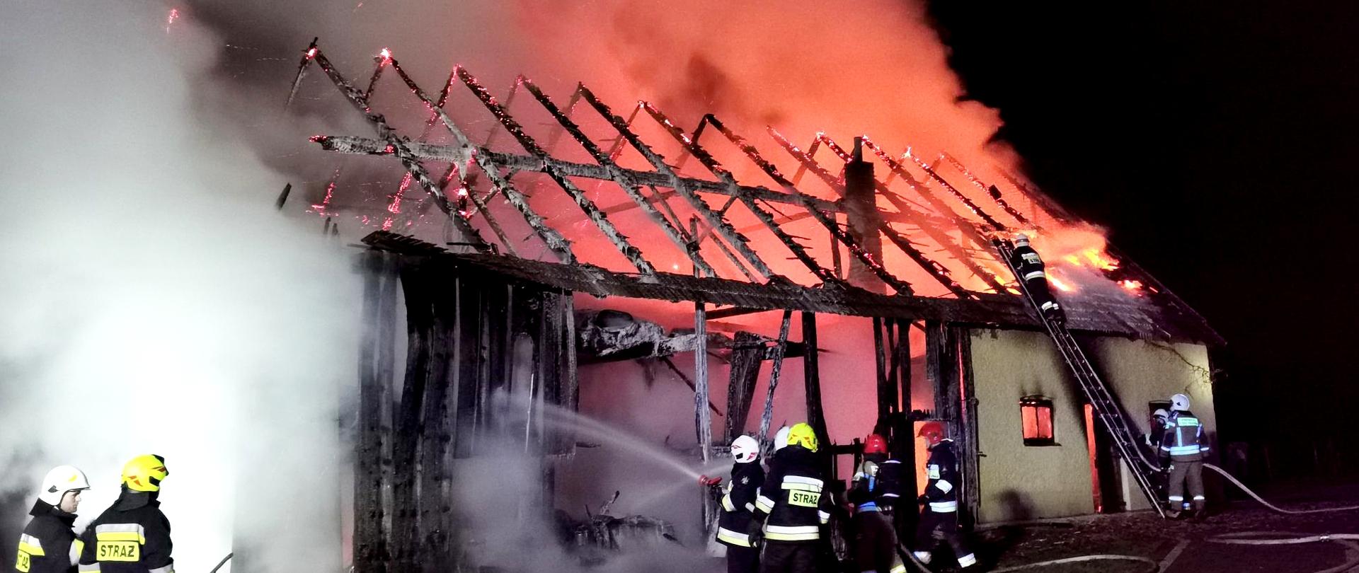 Zdjęcie przedstawia strażaków prowadzących akcję gaszenia pożaru stodoły. Nad całkowicie spalonym dachem widoczne są kłęby gęstego białego dymu, rozświetlone w prawej części fotografii, czerwoną łuną od płomieni buchających ponad powierzchnię budynku. Na pierwszym planie znajdują się trzy dwuosobowe roty strażaków gaszących płonący budynek, w tym jedna rota pracuje z wykorzystaniem drabiny przystawionej do dachu płonącego budynku.