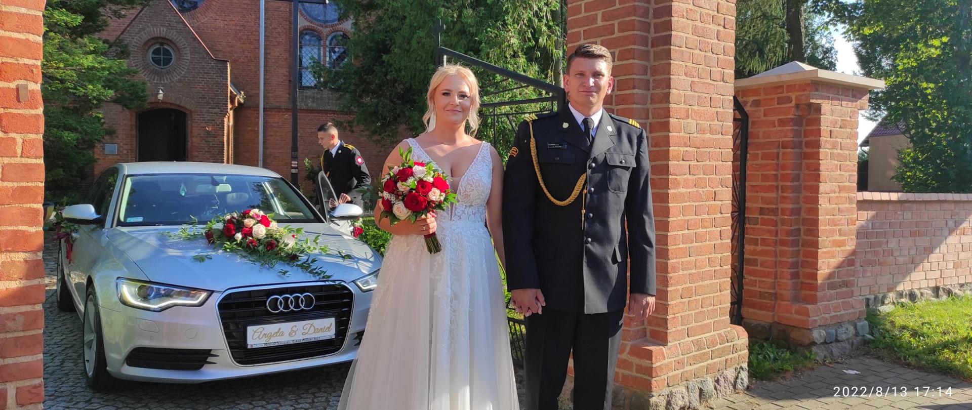 Na zdjęciu strażak w mundurze galowym z małżonką w białej sukni po wyjściu z kościoła. W tle srebrne przystrojone auto oraz kościół z cegły.
