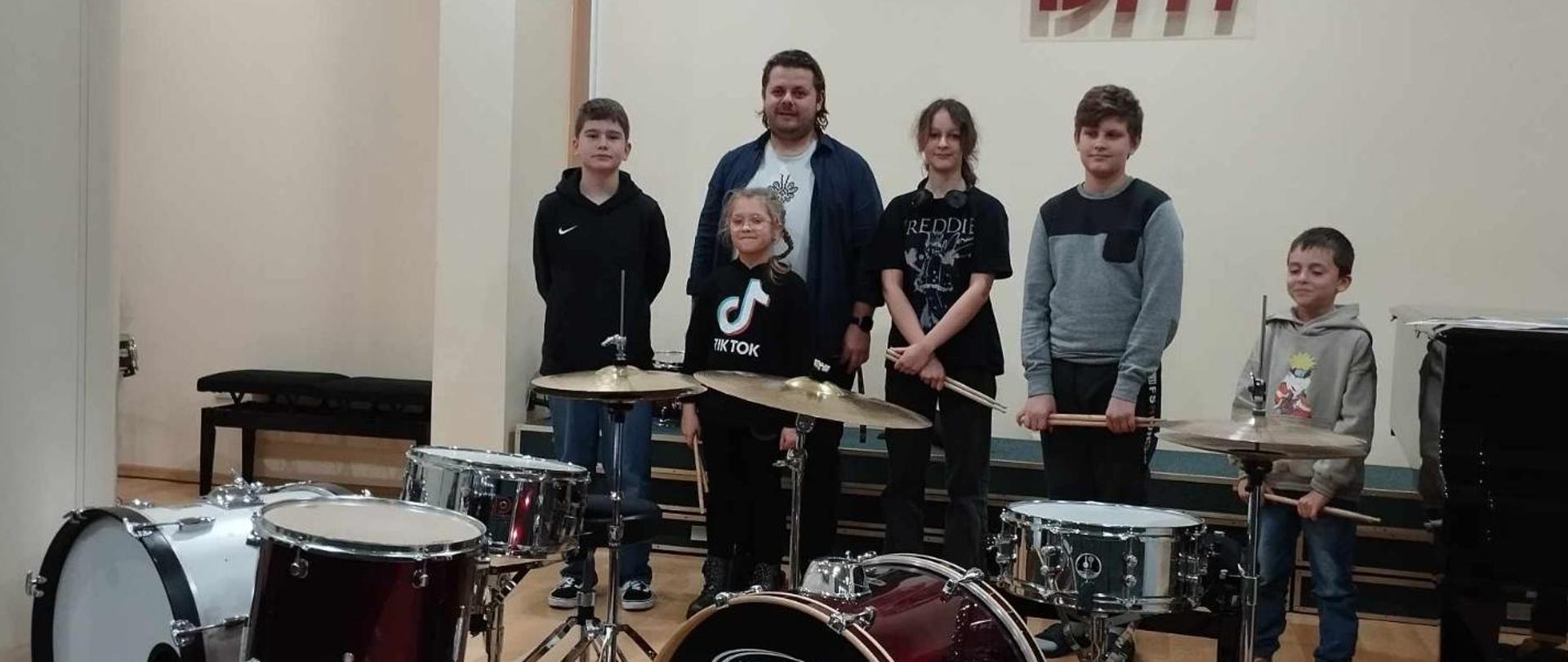 Zdjęcie przedstawia na samym dole zestaw perkusyjny na scenie w sali koncertowej za zestawem perkusyjnym stoją uczniowie i nauczyciel pan Marek Burda.