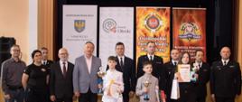 W sali w Miejskim Domu Kultury w Oleśnie są ustawieni w szeregu laureaci II kategorii wiekowej etapu powiatowego OTWP, organizatorzy i zaproszeni goście. W tle stoją 4 banery: Samorządu Województwa Opolskiego, Starostwa Powiatowego, OTWP, Komendy Powiatowej Państwowej Straży Pożarnej w Oleśnie.
