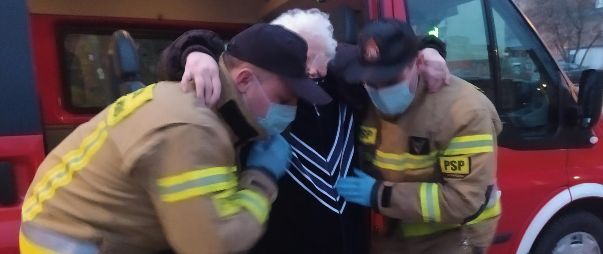 Dwóch strażaków pomaga starszej kobiecie wysiąść z samochodu