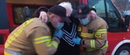 Dwóch strażaków pomaga starszej kobiecie wysiąść z samochodu