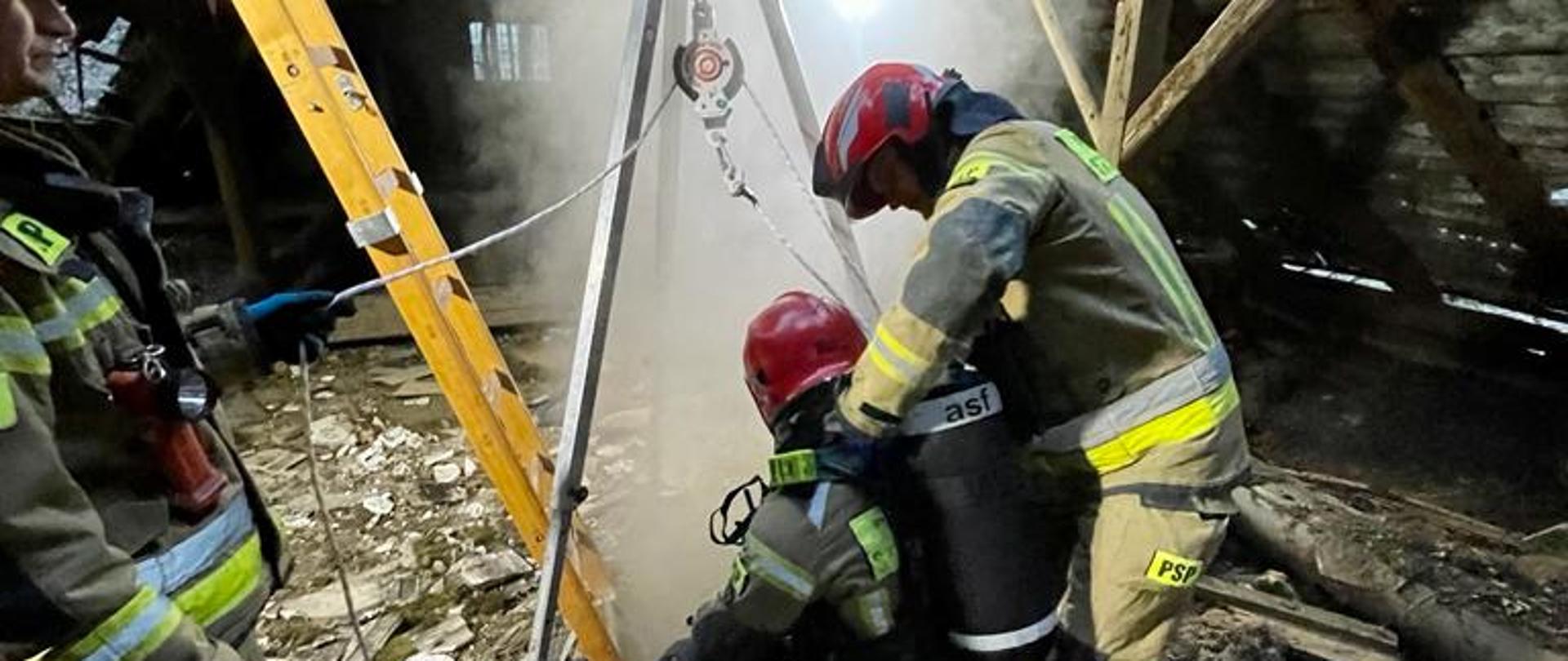 Strażacy obok trójnogu oraz otworu, z którego wydobywa się dym