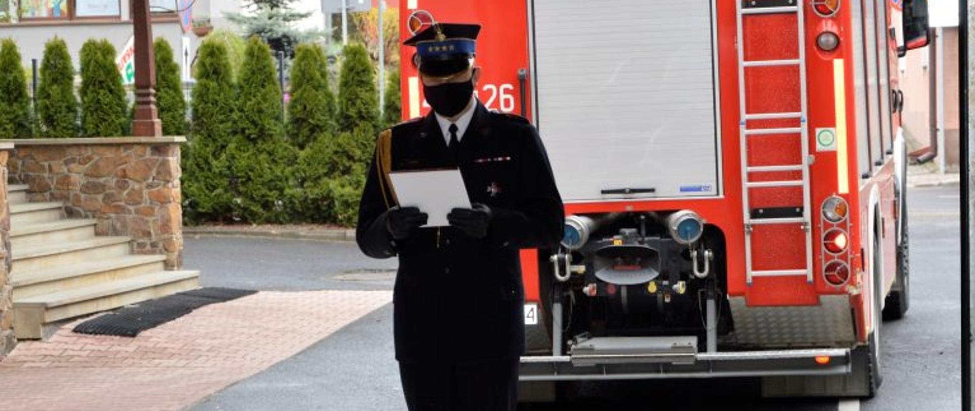 Na zdjęciu komendant powiatowy w umundurowaniu galowym czytający na stojąco list gratulacyjnych strażakom, w tle wejście do komendy oraz samochód pożarniczy