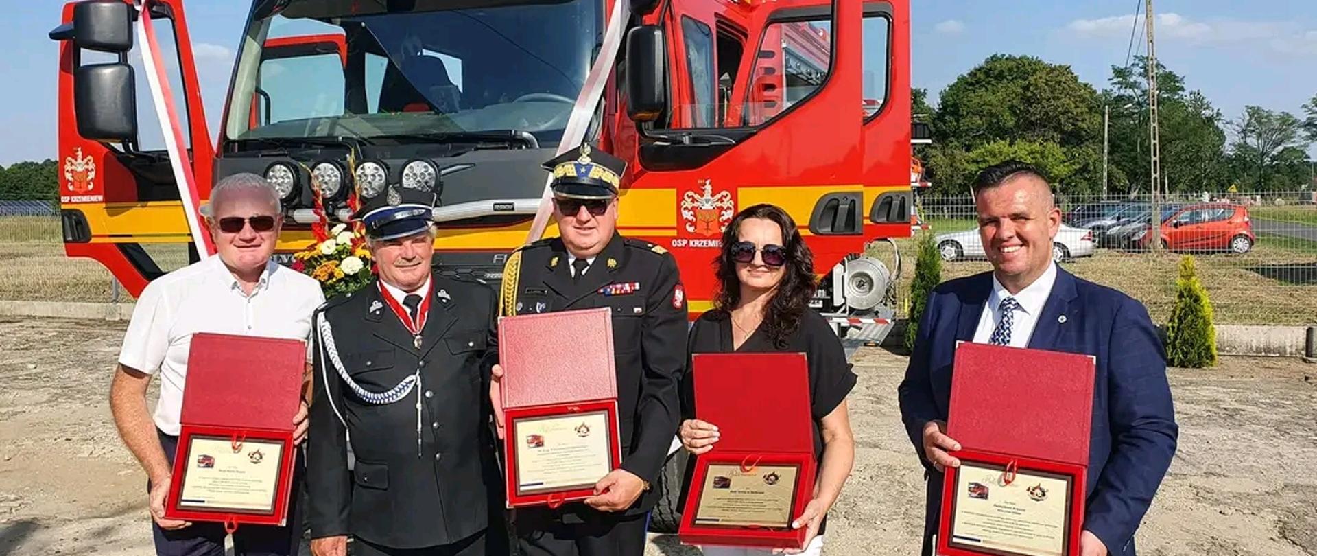 Na tle dużego samochodu strażackiego stoi 5 osób w środku stoi łódzki komendant wojewódzki , po jego lewej stronie pani z rady gminy Dalików obok niej wójt gminy Dalików. Po prawej stronie komendanta stoi przedstawiciel OSP Krzemieniew oraz Poseł na sejm RP Piotr Polak. Trzymają otwarte czerwone pudełka w których pookazują podziękowania 