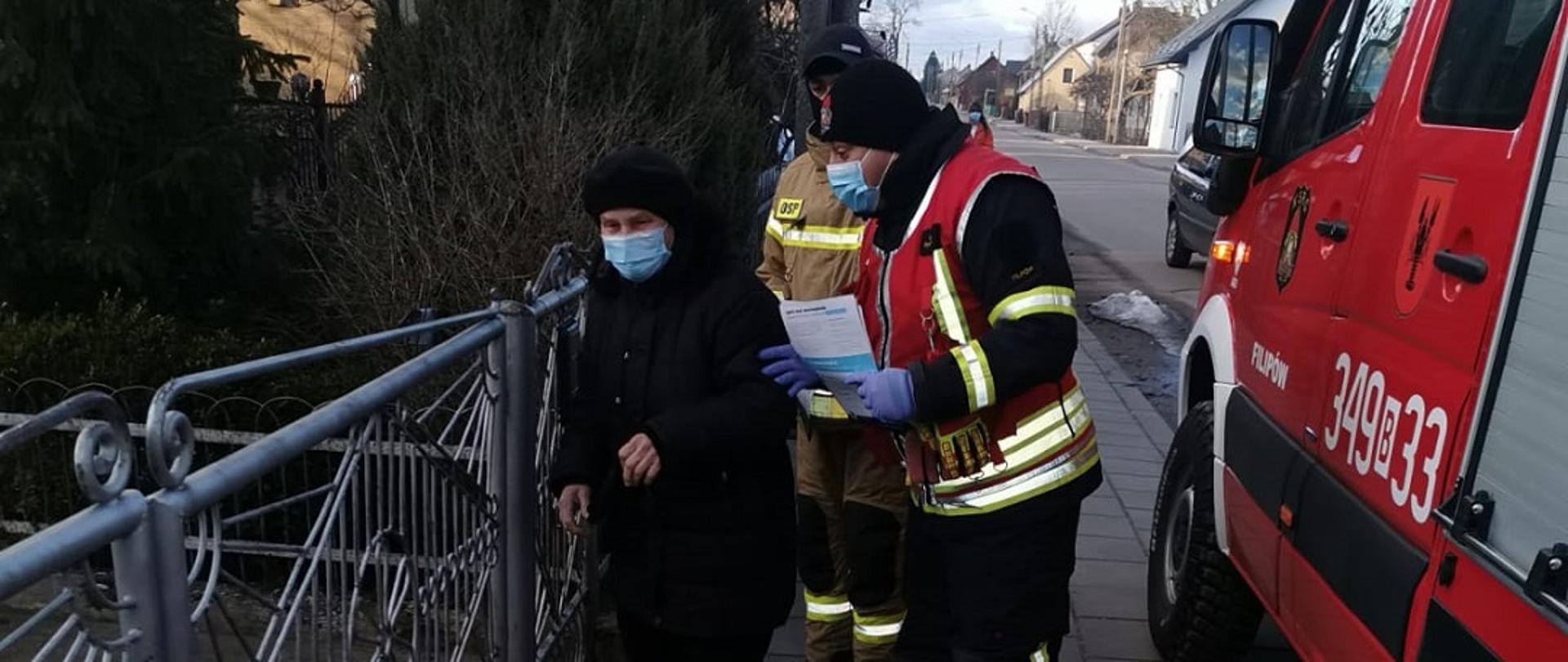 Na zdjęciu widocznych jest dwóch strażaków przekazujących ulotkę starszej kobiecie. Po prawej stronie zdjęcia widoczny jest samochód pożarniczy.