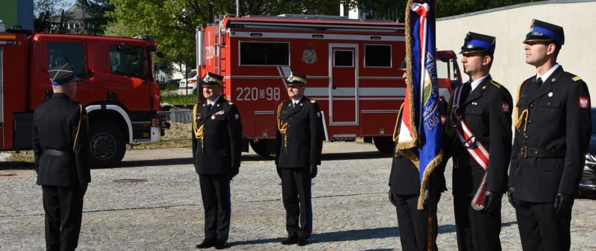 Obraz przedstawia dowódcę uroczystości składającego meldunek Zastępcy Komendanta Głównego. Z prawej strony poczet sztandarowy. W tle samochody pożarnicze. Strażacy w umundurowaniu galowym. Pora dzienna.