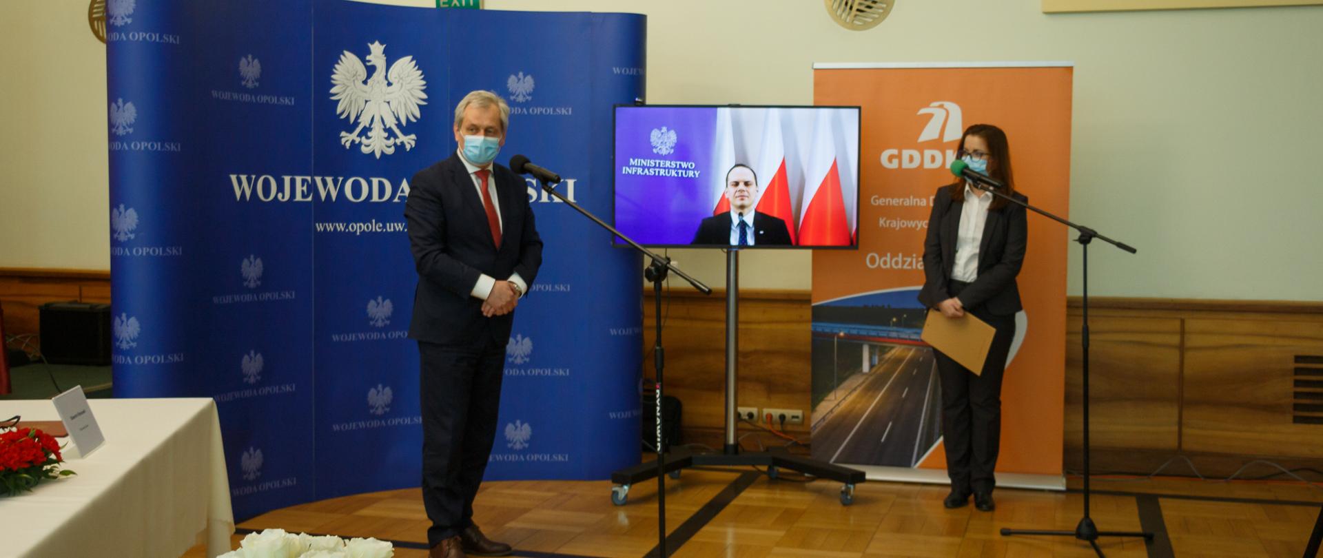 Po lewej stronie kadru widoczny, stojący bokiem do aparatu stół, przepasany białym obrusem. Na nim widać bukiet biało-czerwonych kwiatów oraz plastikową, dużą wizytówkę z imieniem i nazwiskiem wojewody. Na środku kadru widać mikrofon stojący na statywie. Po jego lewej stronie, obrócony bokiem do kadru, wojewoda opolski, który przemawia do zgromadzonych na uroczystości gości. Tuż za mikrofonem na stojaku, widać ekran sporego telewizora na statywie a na nim połączenie online z wiceministrem infrastruktury Weberem. Po lewej stronie monitora, w tle, niebieska ścianka z napisem wojewoda opolski. Po prawej stronie od monitora, także w tle, roll up z napisem GDDKiA. Przed roll upem stoi rzeczniczka wojewody, która przysłuchuje się wypowiedzi wojewody. W dalszej perspektywie, po lewej stronie na ścianie, krzyż. Po prawej stronie na ścianie fragment tabliczki upamiętniający patronat sali, w której odbywa się uroczystość. Po lewej stronie kadru na samym dole widać okazały bukiet biało-czerwonych kwiatów. 