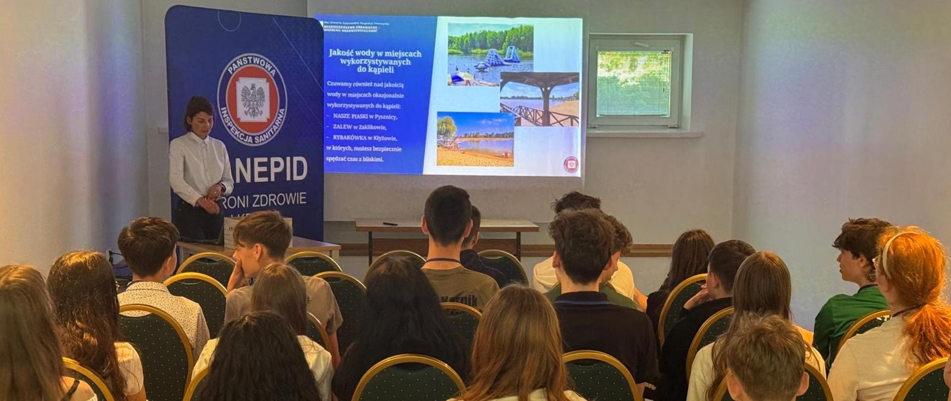Sala wykładowa, na której młodzież siedzi tyłem do obiektywu i słucha prelekcji kobiety stojącej obok ekranu z prezentacją dotyczącą jakości wody w kąpieliskach. W tle baner Państwowej Inspekcji Sanitarnej.
