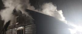 Pożar drewnianego domu letniskowego, z budynku wydobywa się dym. Pora nocna. Strażacy wchodzą do budynku.