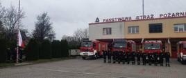 Poczet flagowy wciąga flagę Polski na maszt strażacy stoją przed samochodami gaśniczymi oddając honor.