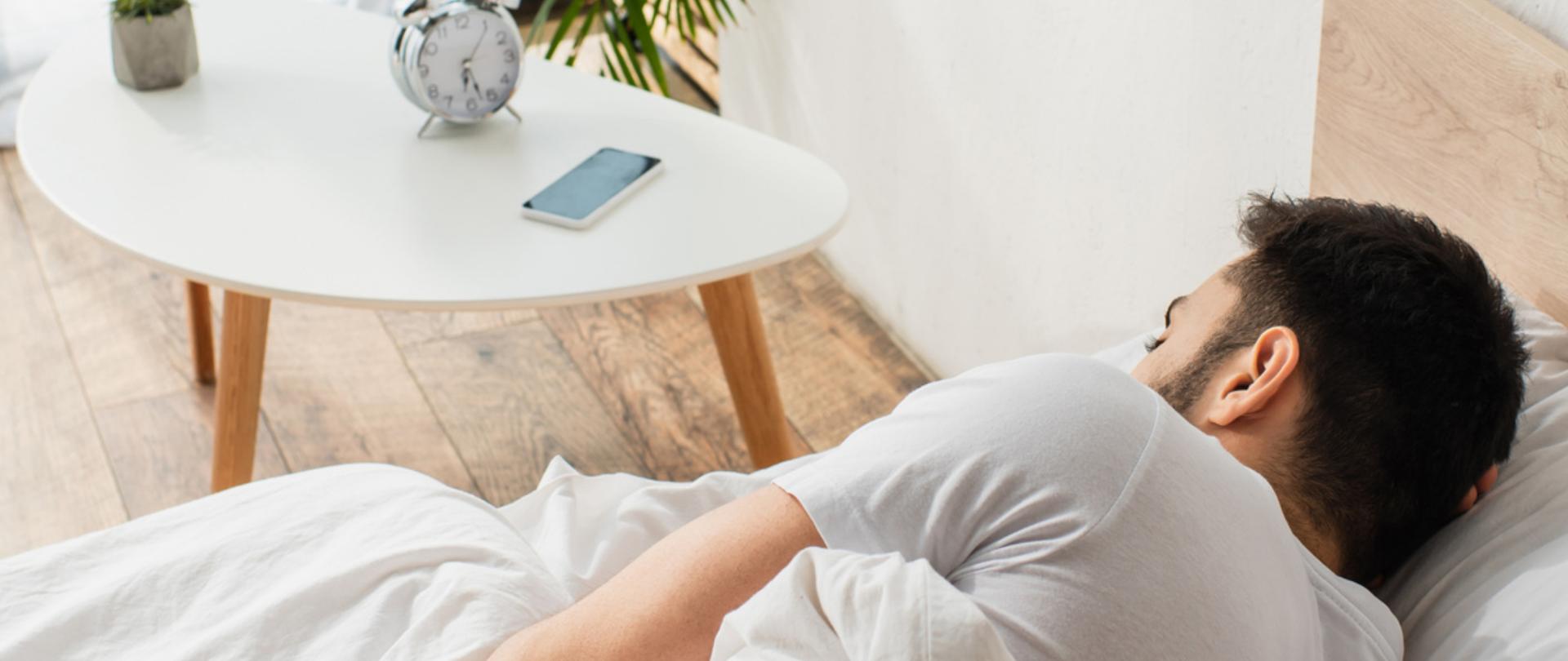 Na zdjęciu jest mężczyzna leżący w łóżku. Obok jest stolik, na którym leży telefon, budzik i kwiat.