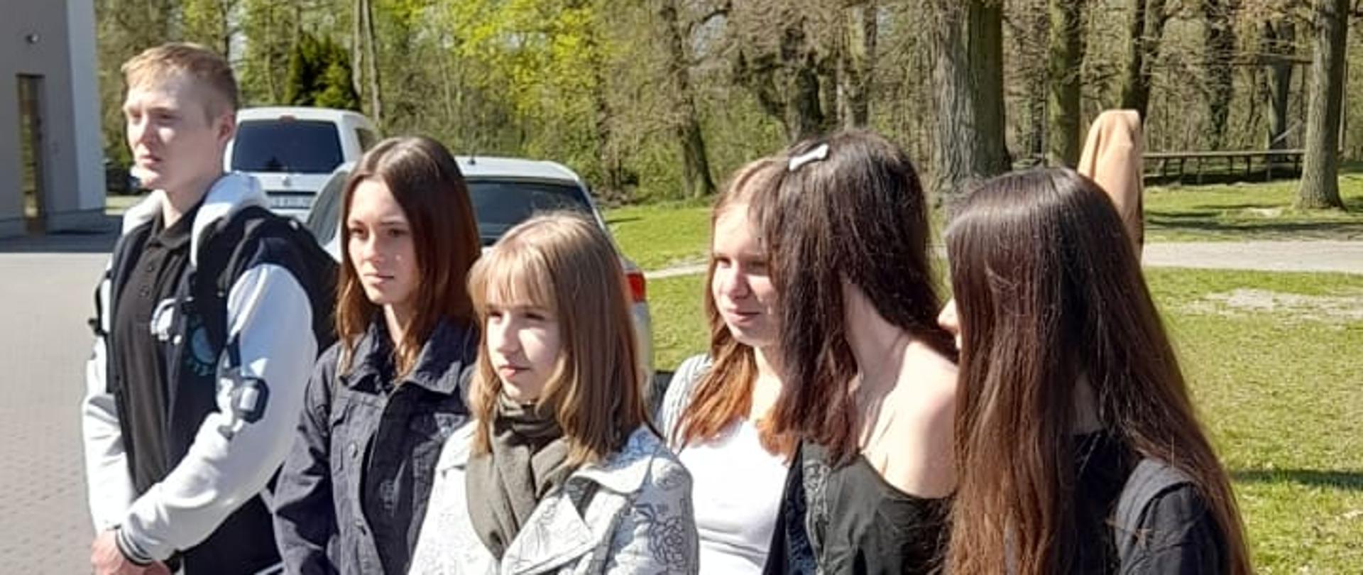 Sześcioro nastolatków stoi w rzędzie na brukowanym chodniku w słoneczny dzień. Od lewej: chłopak w kurtce typu bomber i spodniach bojówkach, dziewczyna w ciemnej dżinsowej kurtce, dziewczyna w jasnym płaszczu we wzory z torebką na ramieniu, dziewczyna w białym T-shircie, dziewczyna w czarnej bluzce i dziewczyna w czarnej koszulce z plecakiem. W tle widać budynek, biały samochód i wysokie drzewa.