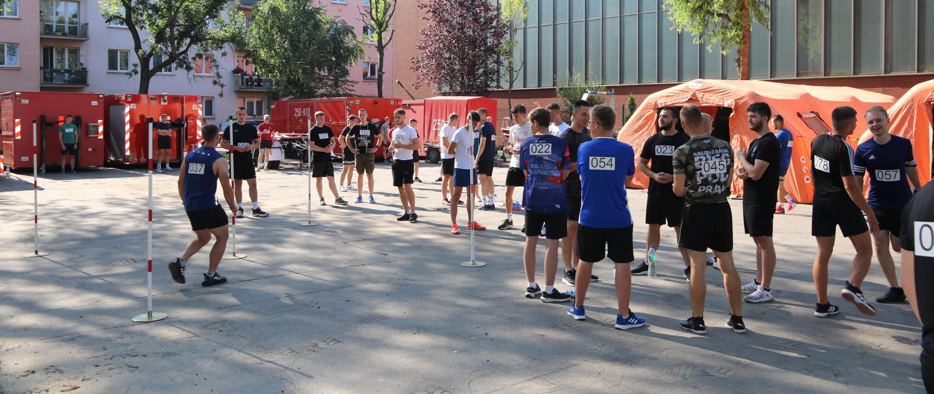 Parking przed halą sportową Szkoły Aspirantów PSP w Krakowie. Na asfaltowej powierzchni rozstawione, pełniące funkcję szatni, pomarańczowe – pneumatyczne – namioty. Przed nimi młodzi ludzie w strojach sportowych i naklejonymi numerami startowymi czekają na swoją kolej. Część dokonuje rozgrzewki. 