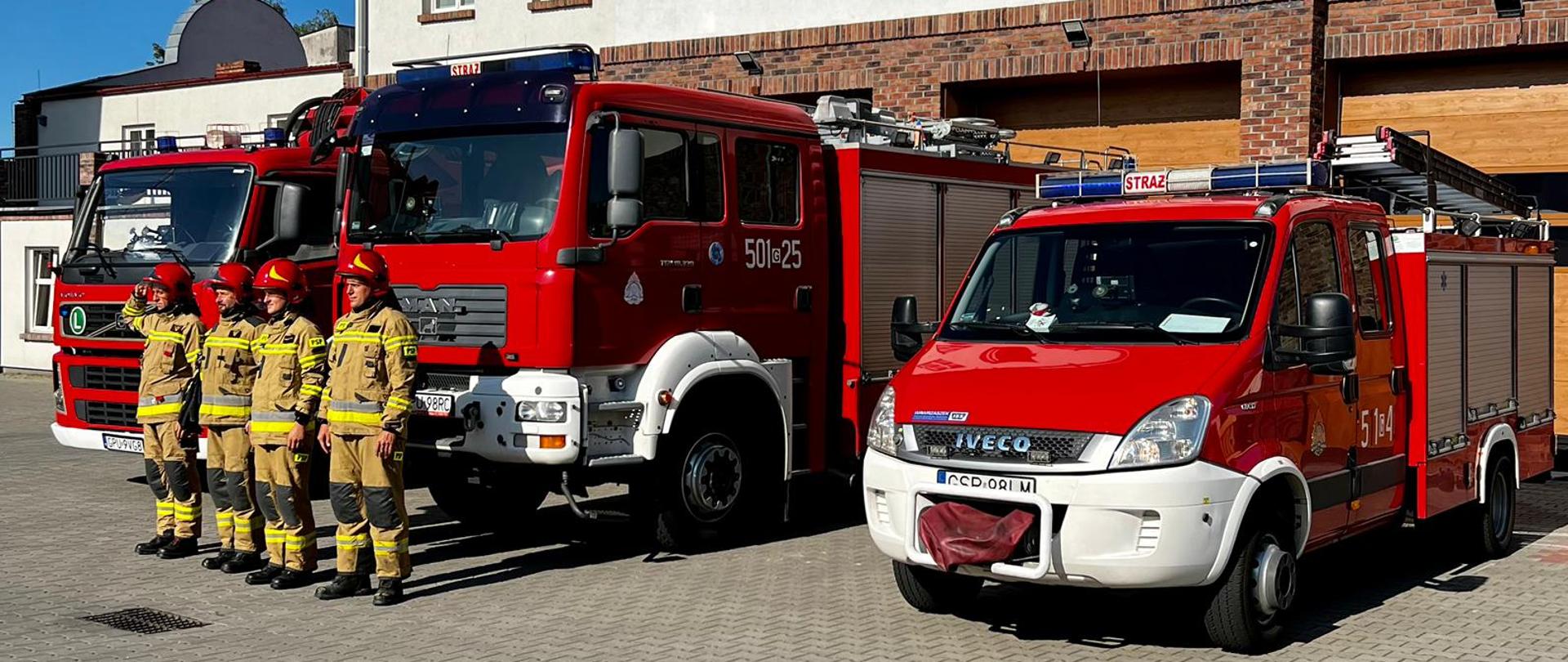 Na zdjęciu strażacy ubrani w ubrania specjalne koloru piaskowego stoją na baczność na tle samochodów strażackich. 