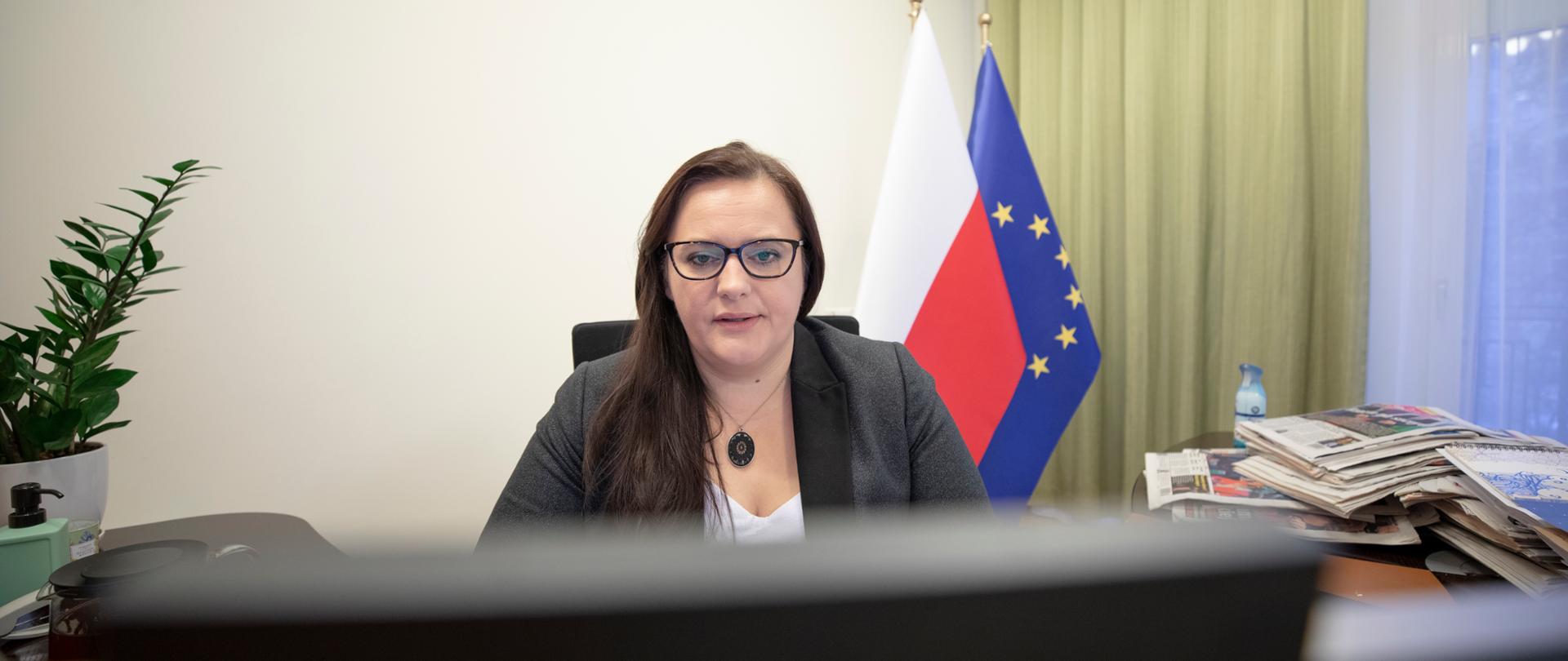 Wiceminister Małgorzata Jarosińska-Jedynak siedzi przed monitorem. Za nią z prawej strony dwie flagi Polski i UE. Z lewej kwiatek w doniczce.