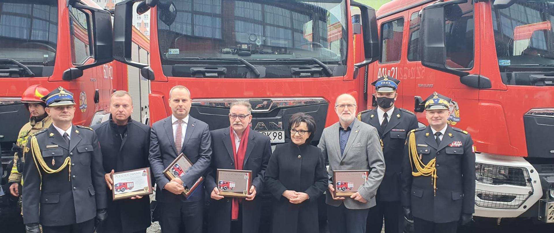 na zdjęciu widać oficjalnych gości uroczystego przekazania nowych pojazdów pożarniczych, w tym między innymi panią Marszałek Elżbietę Witek. Stoją na tle czerwonych samochodów pożarniczych