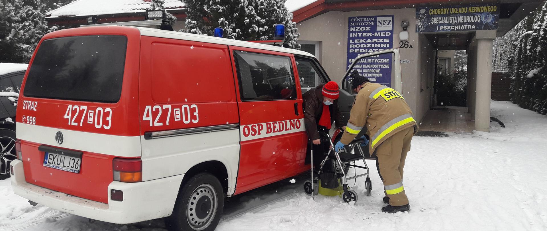Strażak pomagający starszej osobie dotrzeć do punktu szczepień. W tle wóz strażacki OSP.