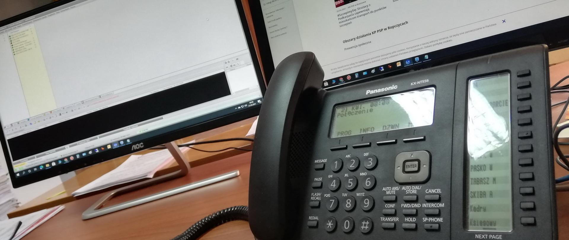 Telefon biurkowy z klawiaturą numeryczna na, postawiony na biurku, w tle monitory komputerowe.