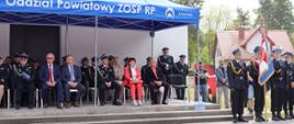 grupa kilkunastu zaproszonych gości siedzi na krzesłach pod namiotem z napisem Oddział Powiatowy ZOSP RP, na pierwszym planie poczet sztandarowy z Komendy Powiatowej PSP w Łowiczu ubrany w mundur wyjściowy z rogatywką na głowie, jedna osoba w poczcie sztandarowym trzyma sztandar komendy w dłoni opartej drzewcem o kostkę brukową