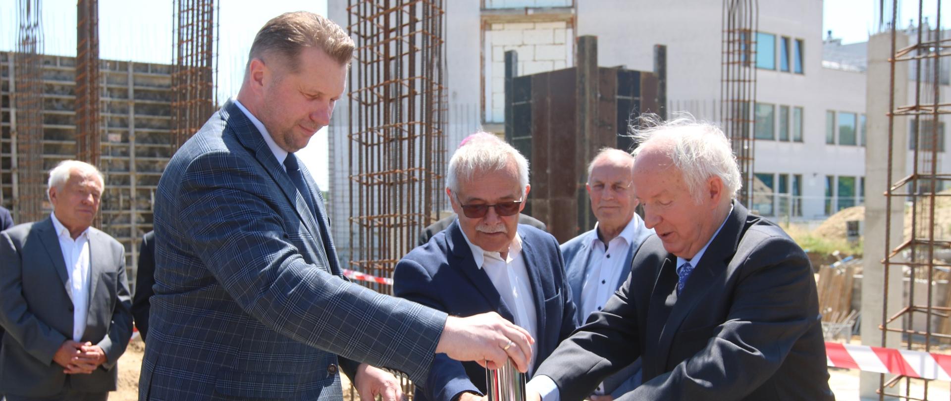 Minister Czarnek i dwóch mężczyzn w garniturach wkładają metalowy walec do betonowego czworokąta.