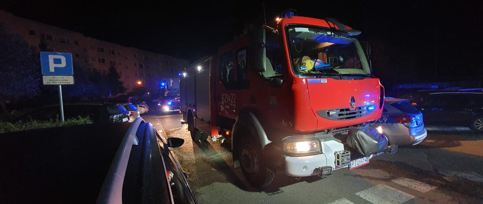 Na drodze przy bloku stoją samochody pożarnicze uczestniczące w akcji gaśniczej.