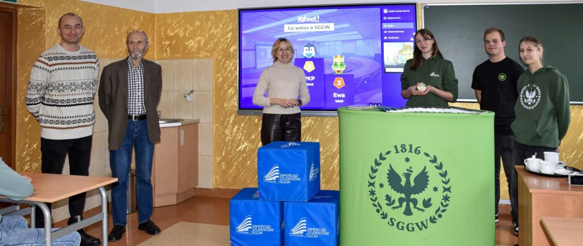 Studenci w strojach z logo SGGW stoją za stoiskiem oznaczonym logo uczelni, za nimi ekran z wyśwtetloną prezentacją promocyjną. Wokół gażety promocyjne uczelni. Obok nauczyciele ZS CKR w Starym Lubiejewie