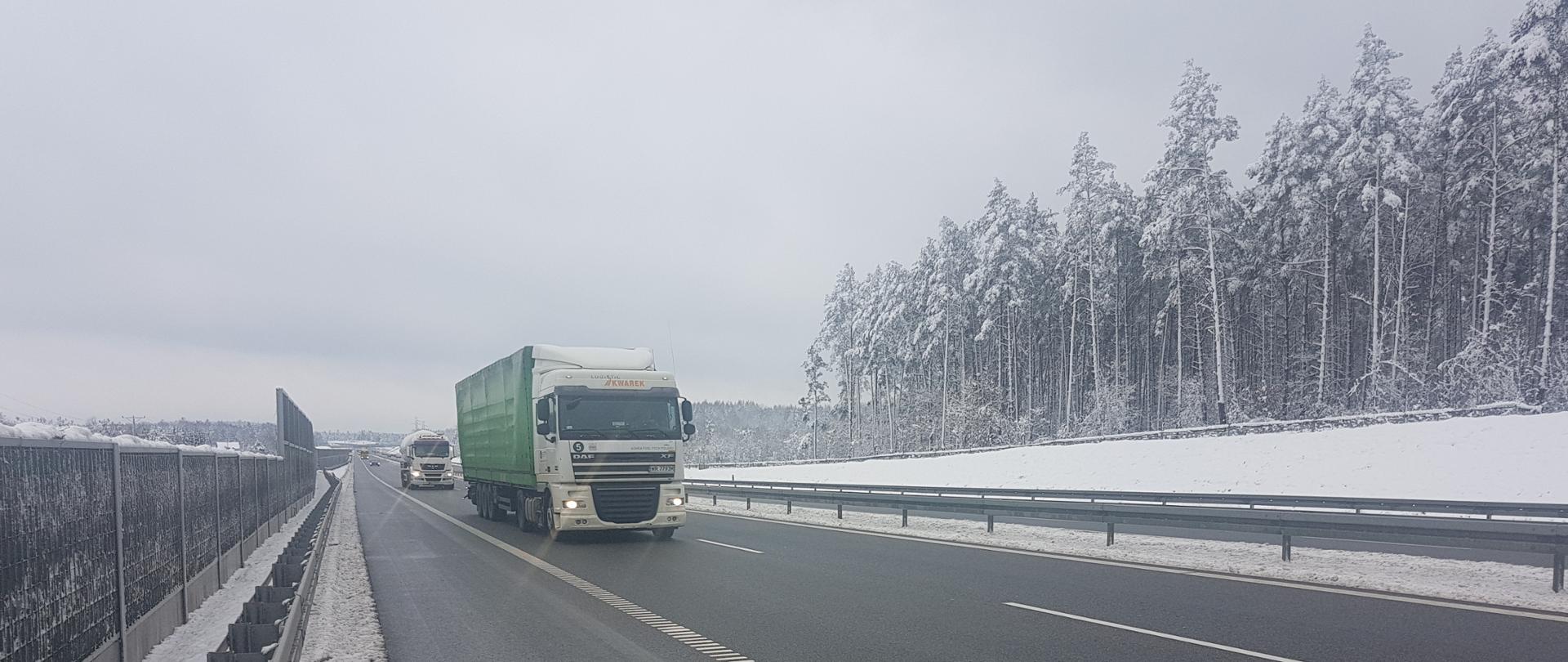 Droga ekspresowa zimą - odśnieżona jezdnia, jadące samochody ciężarowe jeden za drugim, przy drodze bariery i ekrany akustyczne, za nimi ośnieżone na biało drzewa 