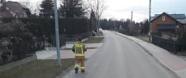 Na zdjęciu widnieje strażak poruszajacy się asfaltową drogą wiejską w ubraniu specjanym strażaka, który dostarcza ulotki pod kolejne adresy. 