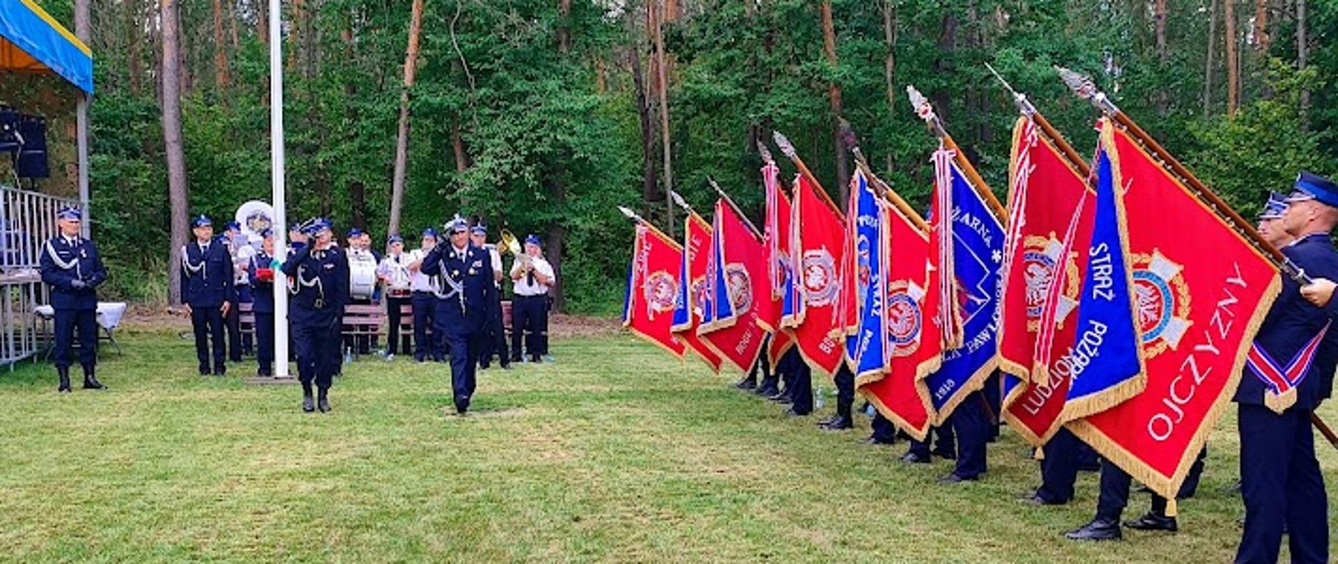 Szeroki plan przedstawiający uroczysty apel na świeżym powietrzu. Na trawiastym placu, na tle lasu, stoi szereg czerwonych sztandarów strażackich ze złotymi haftami i orłami. Po lewej stronie, w stronę sztandarów, maszeruje dwóch oficerów w galowych mundurach, salutując. Pozostali strażacy i goście stoją za nimi. Widoczna jest również scena z żółto-niebieskim dachem.