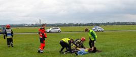 Na zdjęciu widzimy strażaków podczas ćwiczeń na Lotnisku Rudniki. Strażak w umundurowaniu bojowym pomaga osobą, które udają poszkodowanych. Na zdjęciu widoczny jest również Ratownik Medyczny oraz pracownicy lotniska. 