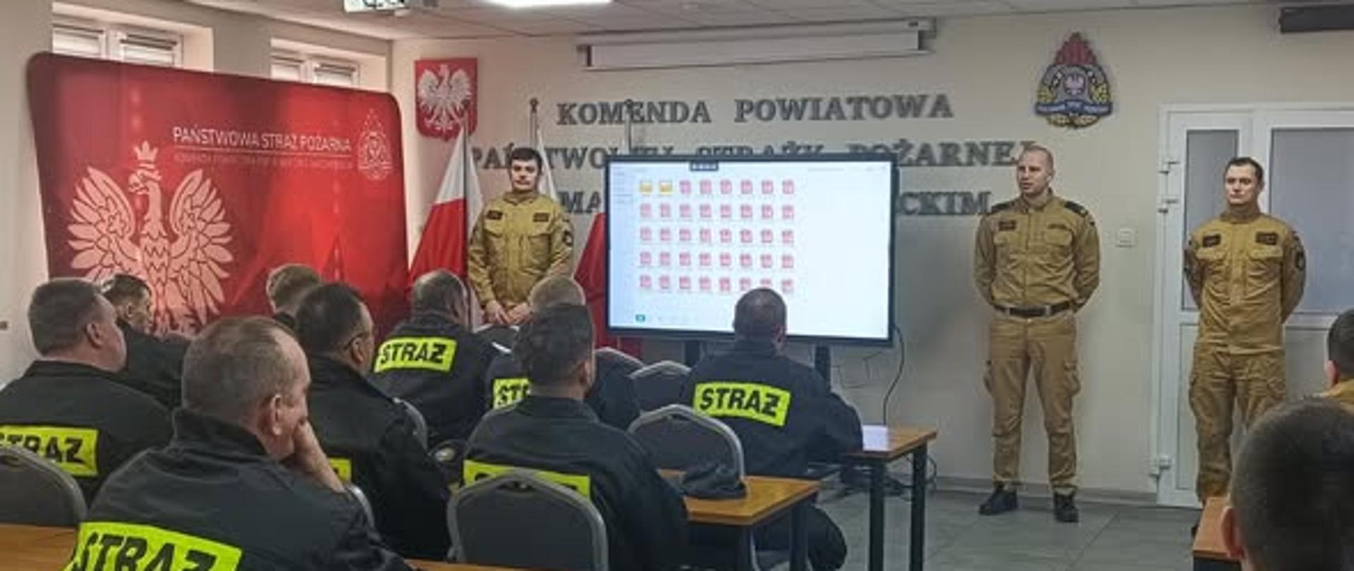 Komendant Powiatowy wraz z dwoma funkcjonariuszami, stoją przy drzwiach i tablicy interaktywnej; rozpoczynają szkolenie. Ubrani są w mundury w kolorze piaskowym. Ochotnicy siedzą w ławkach, ubrani w czarne stroje z napisem STRAŻ na plecach. 