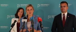 Kobieta w niebieskim garniturze przemawia do mikrofonów na tle ścianki z napisem Reforma26 Kompas Jutra. Obok stoją druga kobieta oraz mężczyzna w garniturze