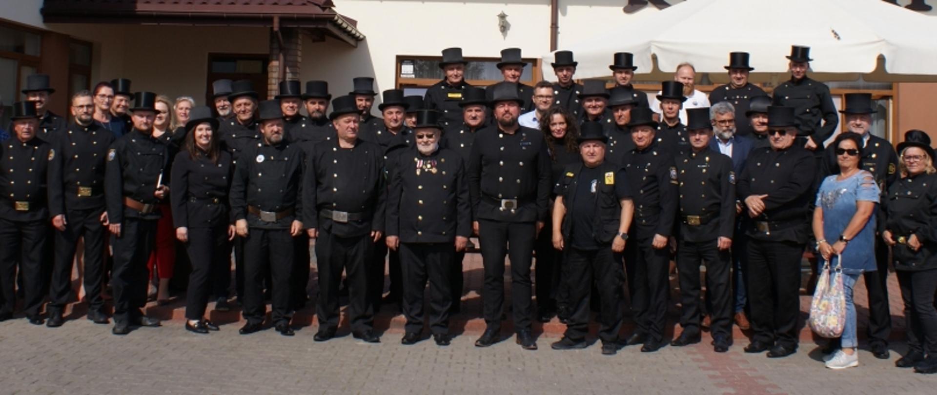 Duża grupa osób, głównie w czarnych mundurach i cylindrach, pozująca do zdjęcia przed budynkiem z oknami i wejściem.